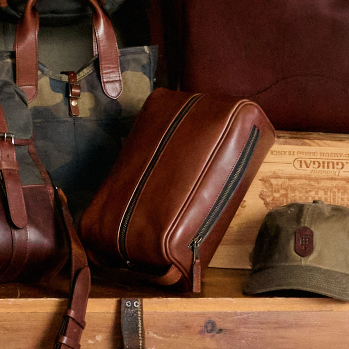 Tom Beckbe Leather Dopp Kit, green camo backpack, and baseball cap rest on a wooden surface.