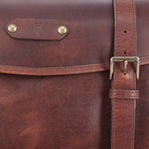 Close-up of a brown leather satchel with brass hardware and a visible embossed logo on the flap.