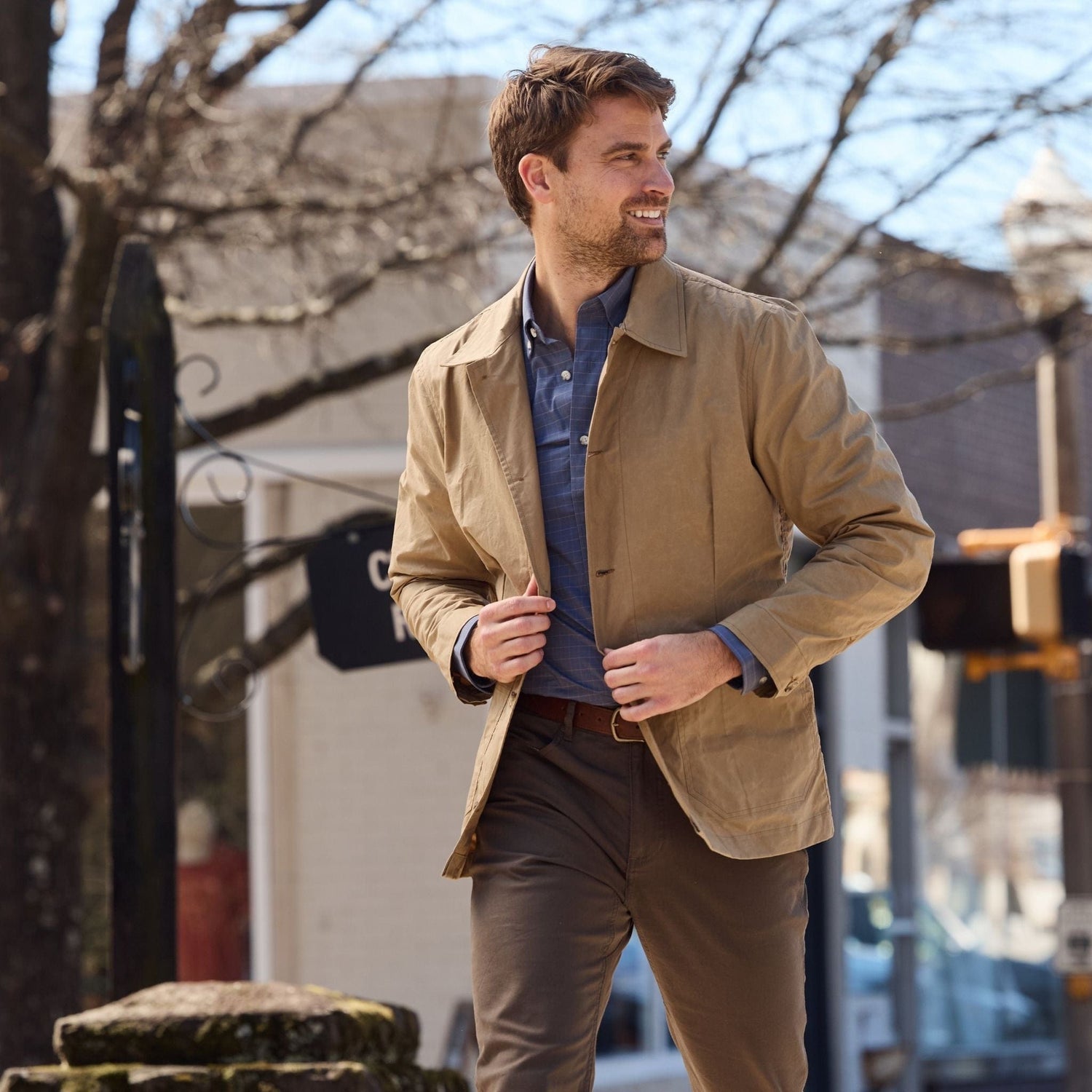 Man strolling outdoors in a Tom Beckbe Livingston Jacket and brown pants, with buildings and bare trees behind him.