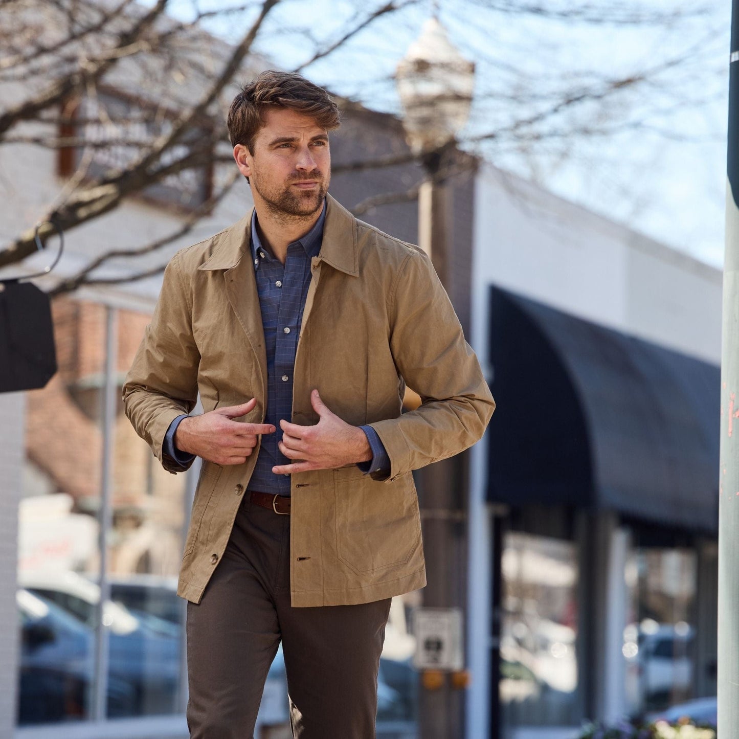 Man walking on a city street in a Tom Beckbe Livingston Jacket and blue shirt, with leafless trees and storefronts behind him.