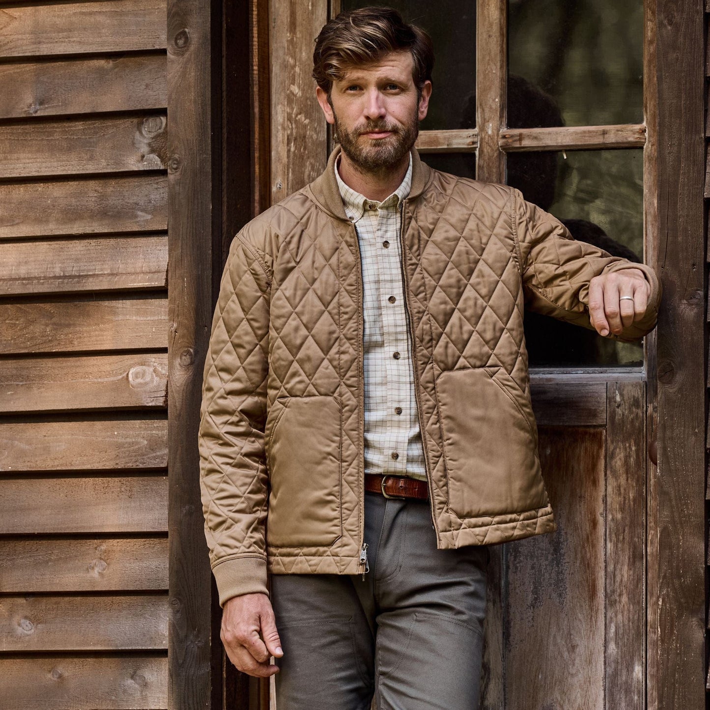Man in vintage style, wearing a Logan Bomber Jacket by Tom Beckbe, leaning against a wooden buildings doorframe.