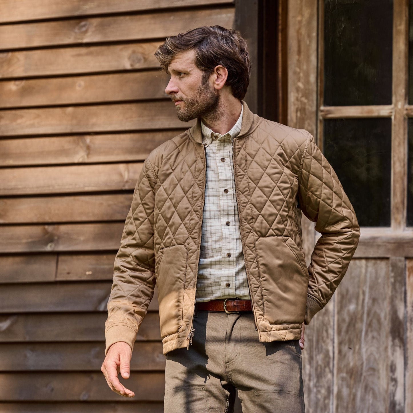 A man in a Logan Bomber Jacket by Tom Beckbe, with a checked shirt, stands beside a wooden building looking sideways.