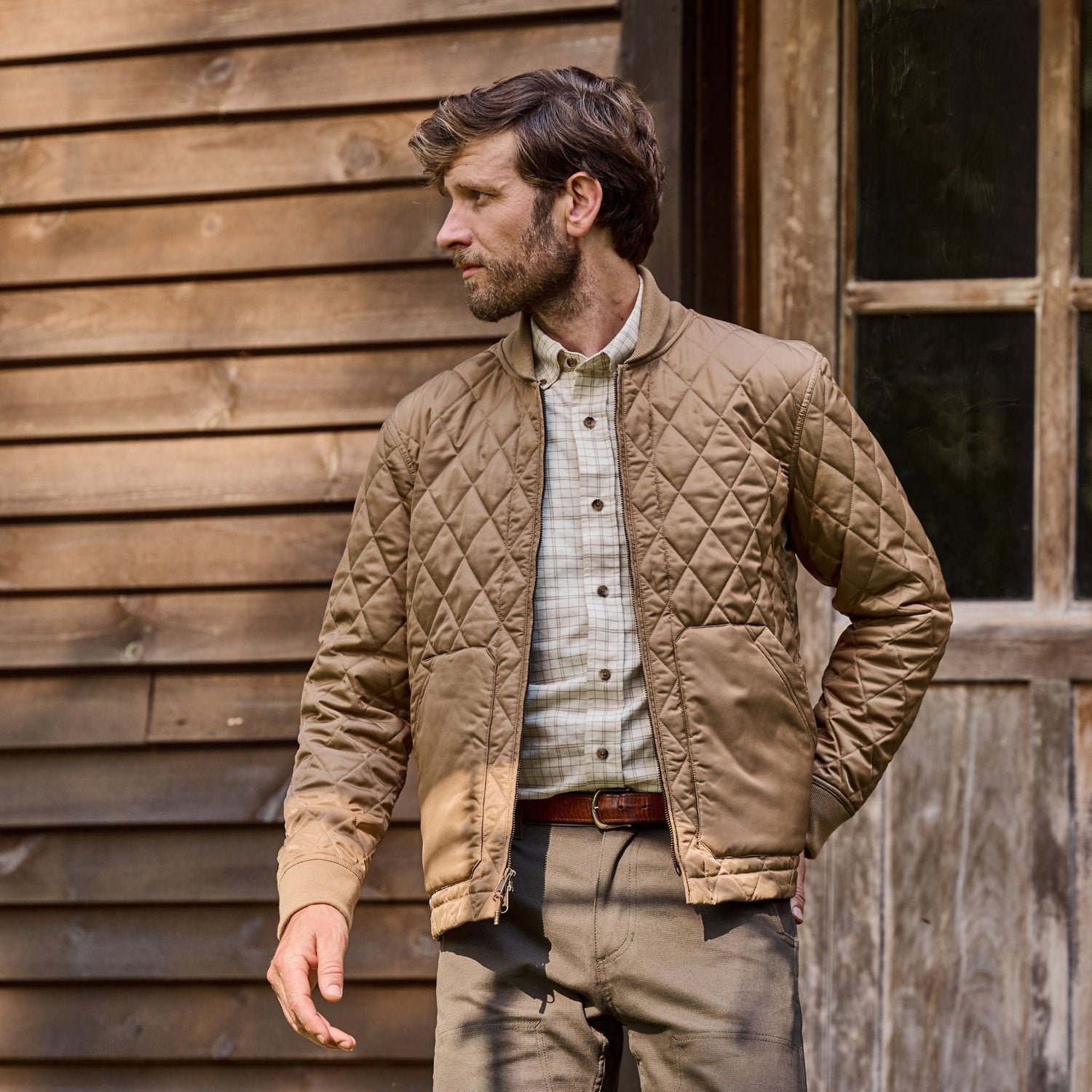 A man in a Logan Bomber Jacket by Tom Beckbe, with a checked shirt, stands beside a wooden building looking sideways.