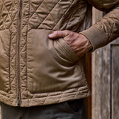 A person in a quilted brown Tom Beckbe Logan Bomber Jacket stands against a vintage wooden background, hand in pocket.