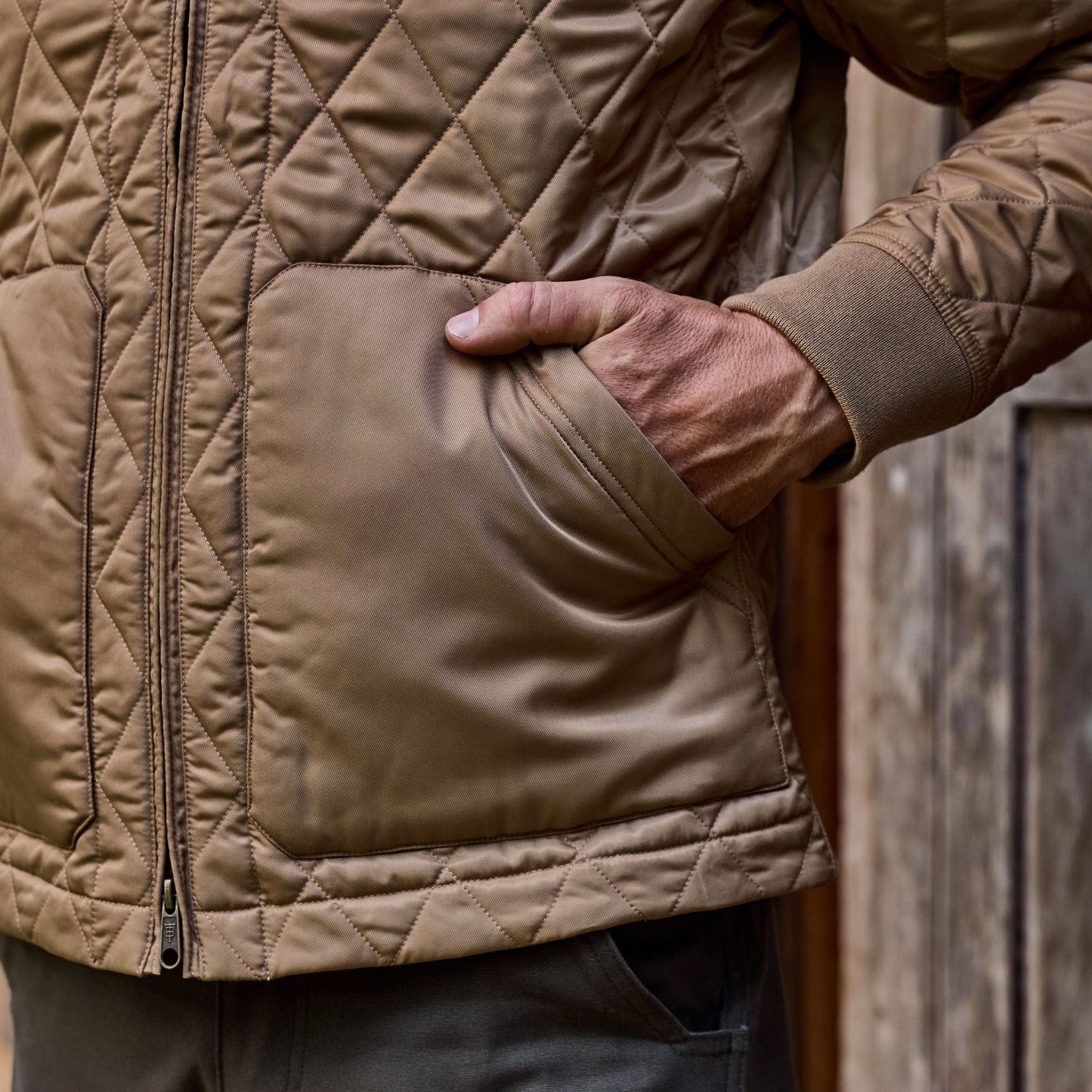 A person in a quilted brown Tom Beckbe Logan Bomber Jacket stands against a vintage wooden background, hand in pocket.