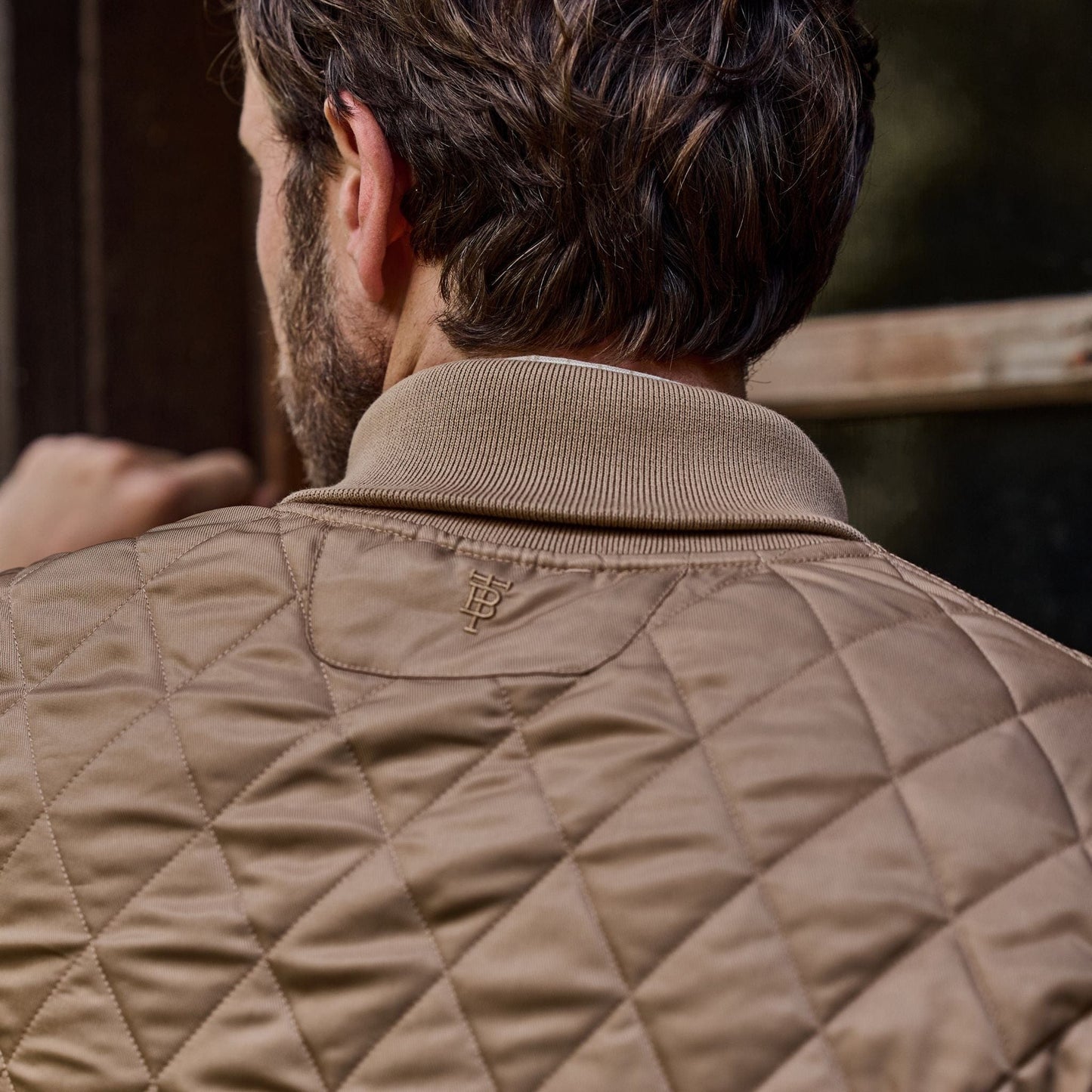 A person in a vintage-style Tom Beckbe quilted brown Logan Bomber Jacket with an embroidered logo, standing by a window.