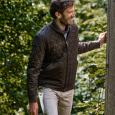 Man in a vintage-style Tom Beckbe Logan Bomber Jacket and beige pants smiles near wooden structure with greenery.