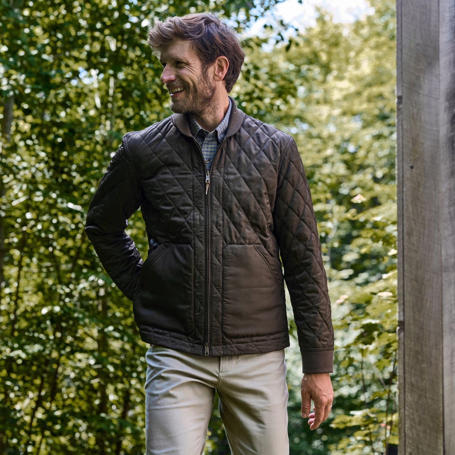 A man in a Tom Beckbe Logan Bomber Jacket and light pants smiles outdoors, hands in pockets, among trees.