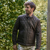 Man in a Tom Beckbe Logan Bomber Jacket stands outside, quilted vintage-style brown with green trees behind him.