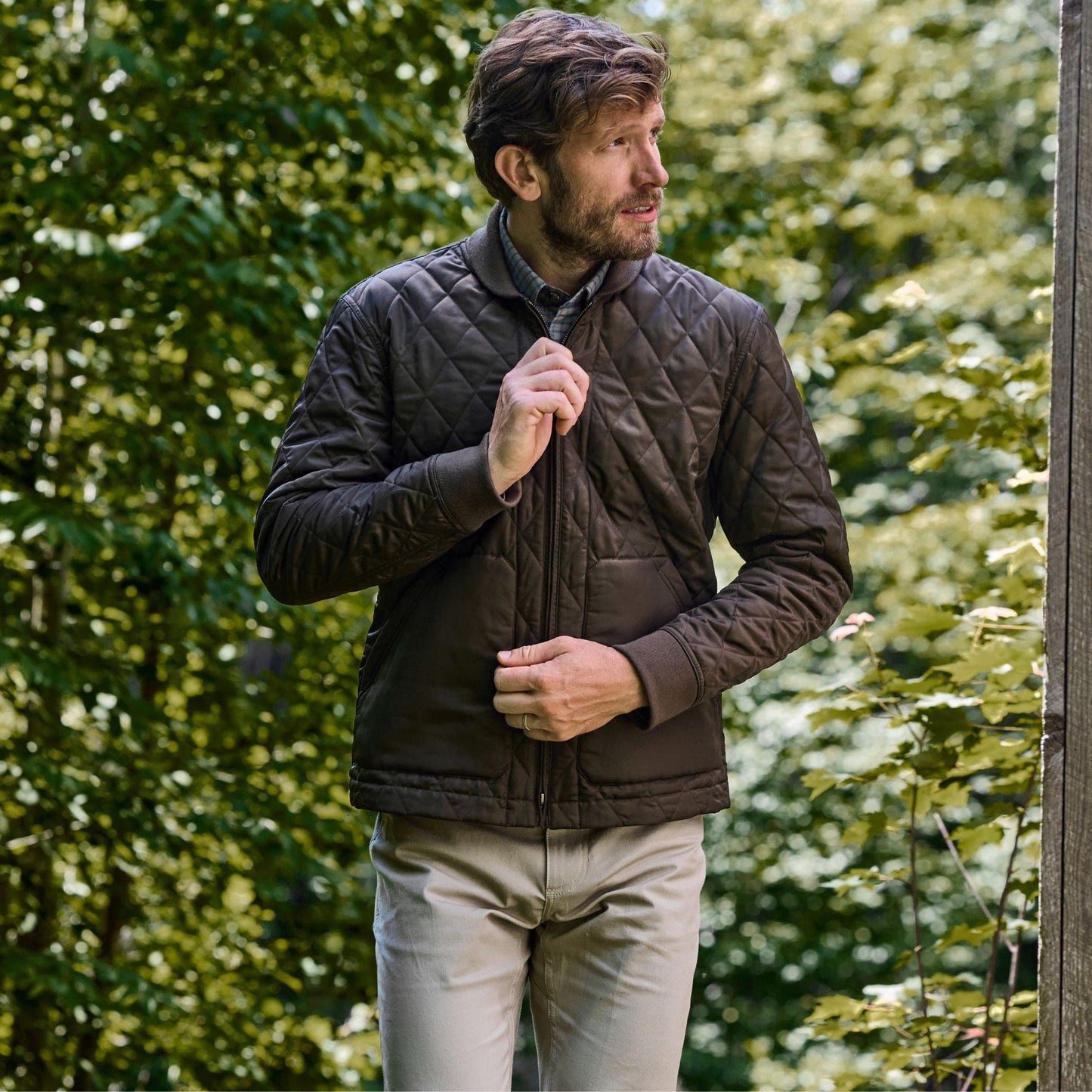 Man in Tom Beckbes Logan Bomber Jacket adjusts collar, embracing vintage style among green foliage.