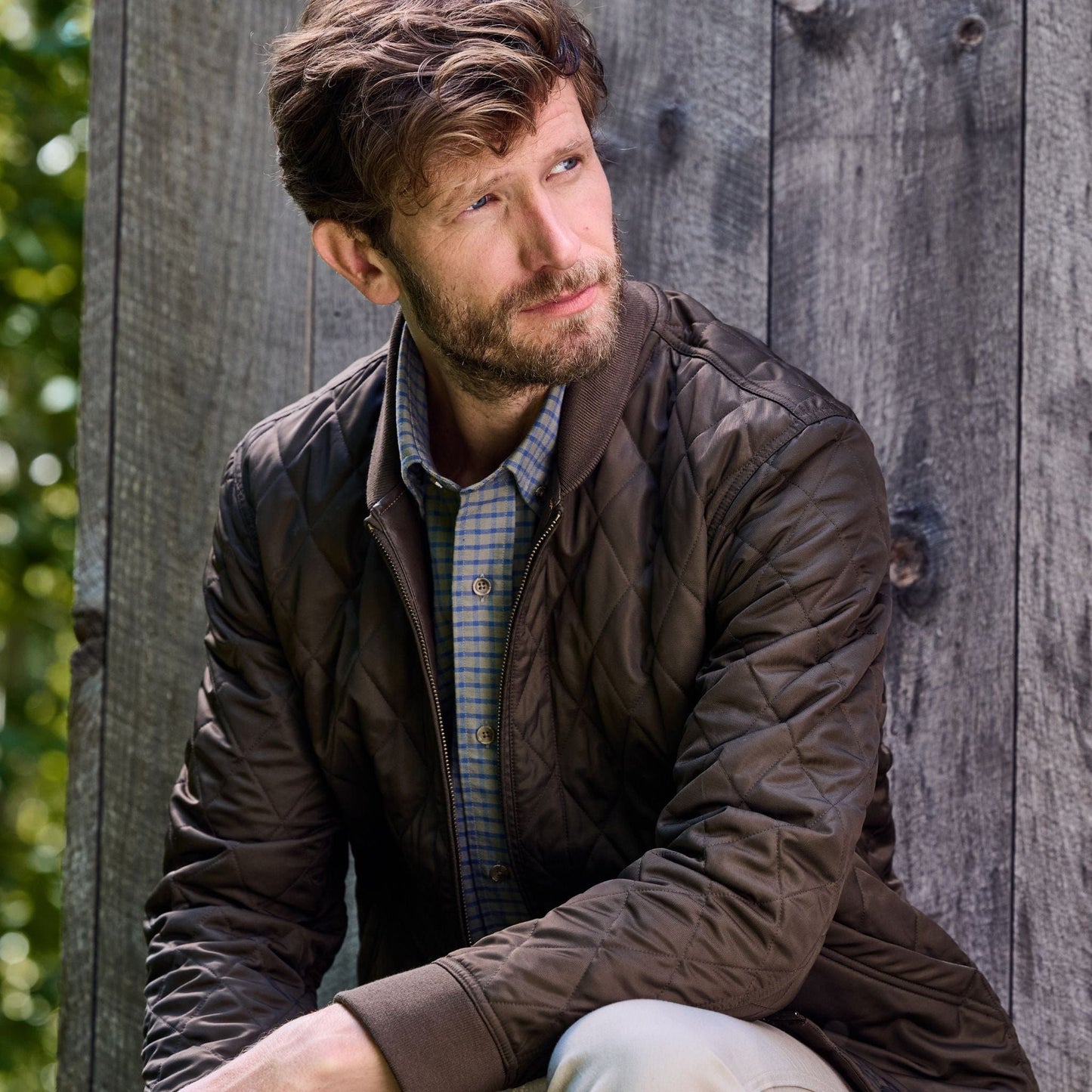 Man in a brown Tom Beckbe Logan Bomber Jacket and plaid shirt, sitting against a wooden wall, looking to the side.