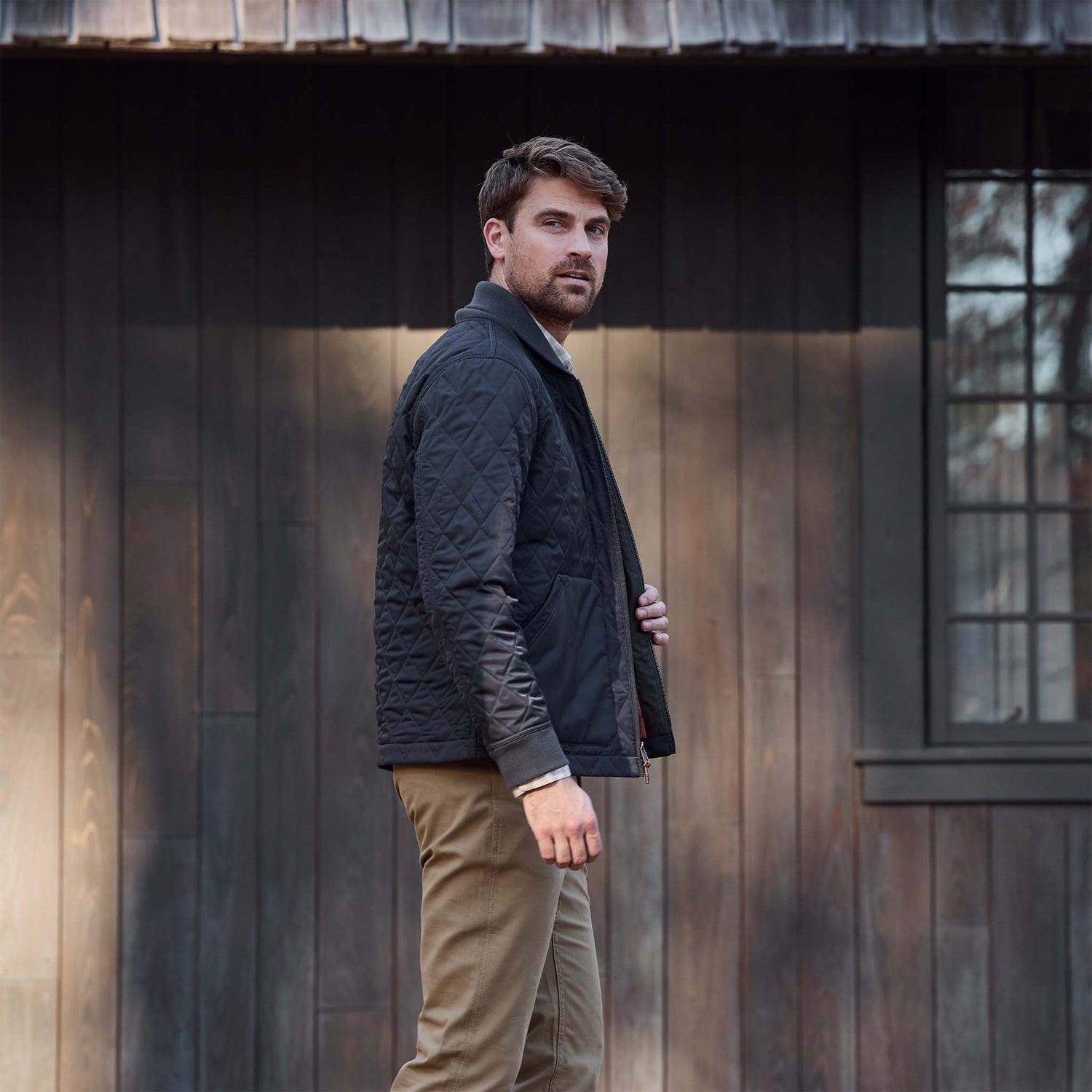 A man in a Tom Beckbe Logan Bomber Jacket and khaki pants stands outside by a wooden building, facing the camera.
