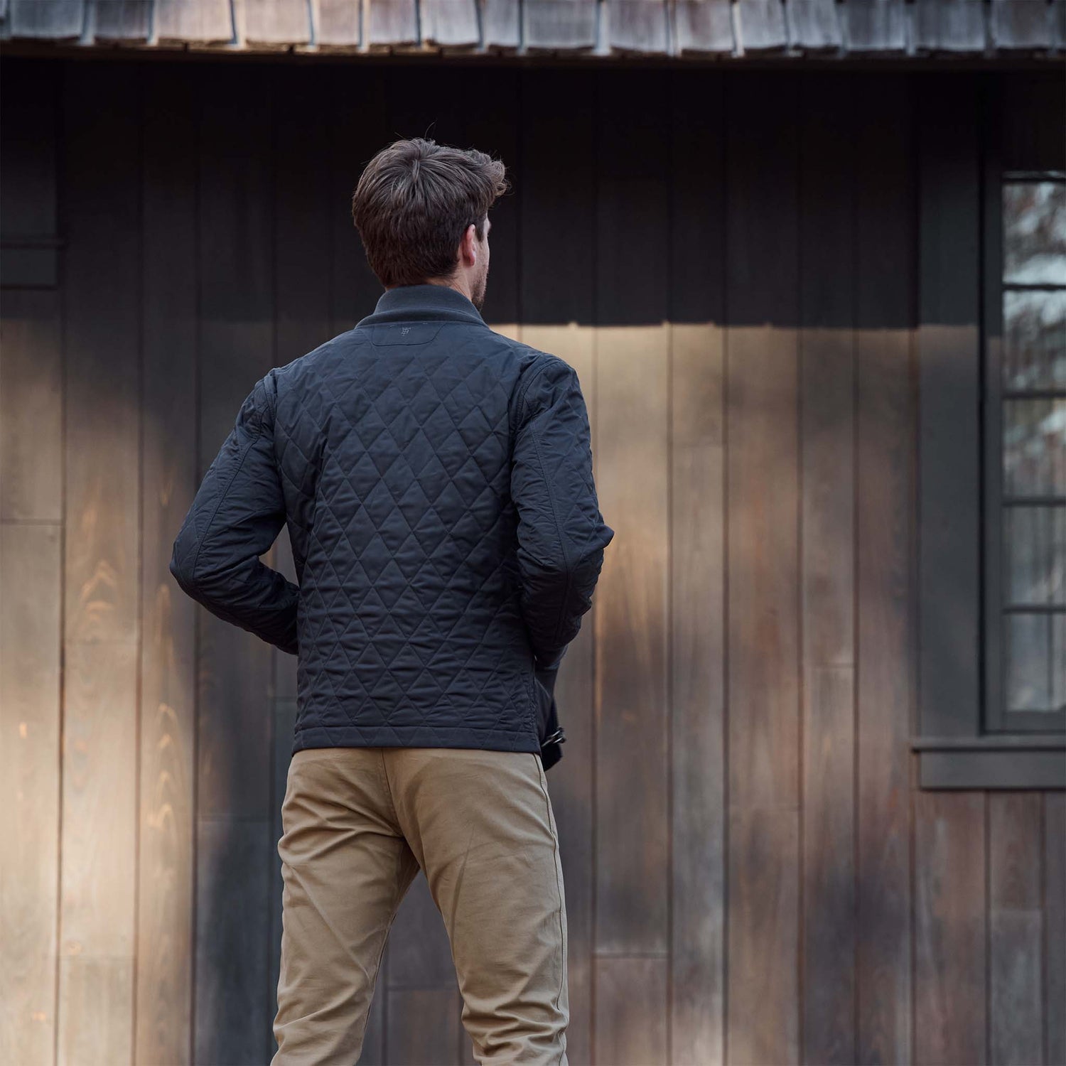 A man in a Tom Beckbe Logan Bomber Jacket and khaki pants stands facing a dark wooden building.
