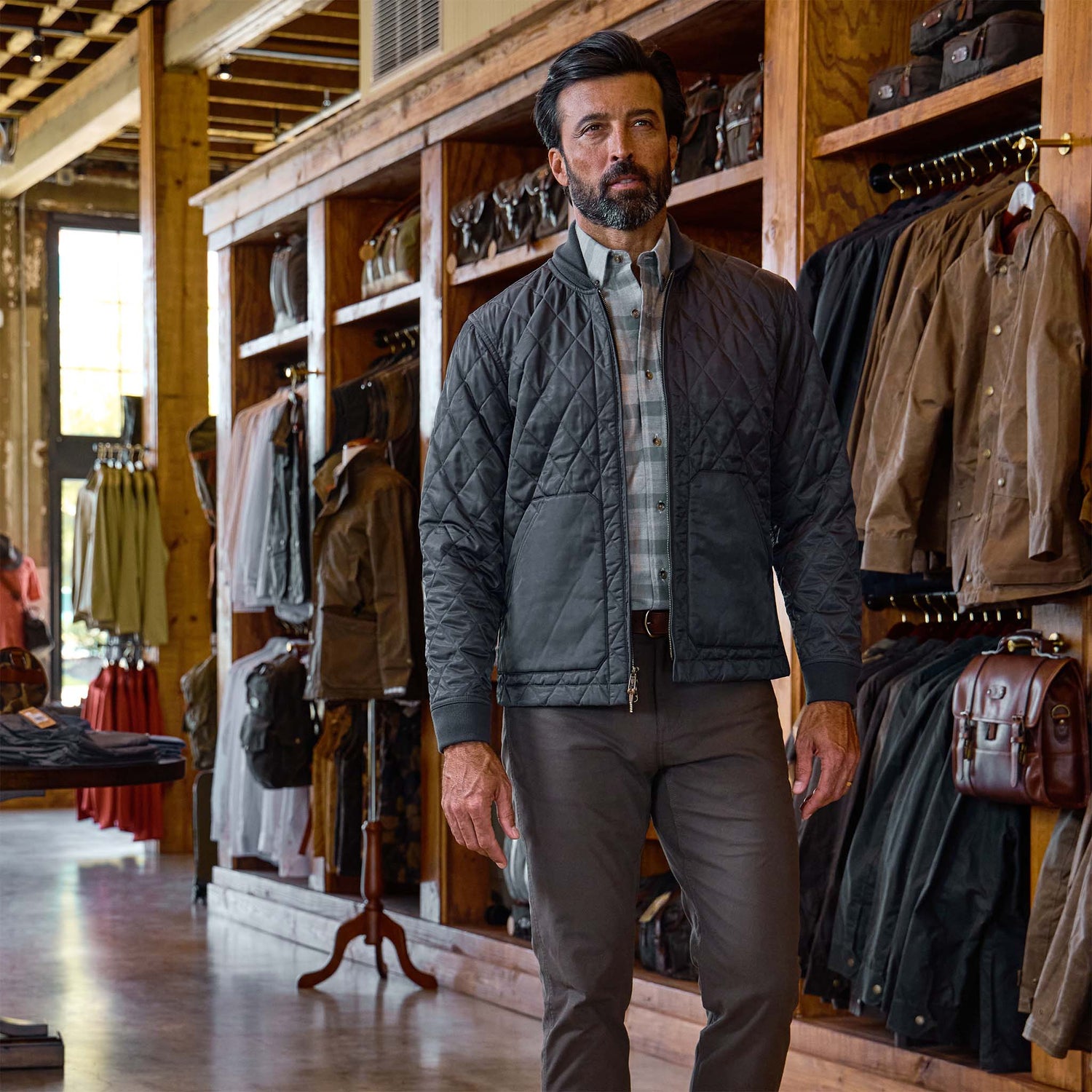 A man in a Tom Beckbe Logan Bomber Jacket and plaid shirt walks through a store with wooden shelves and hanging jackets.