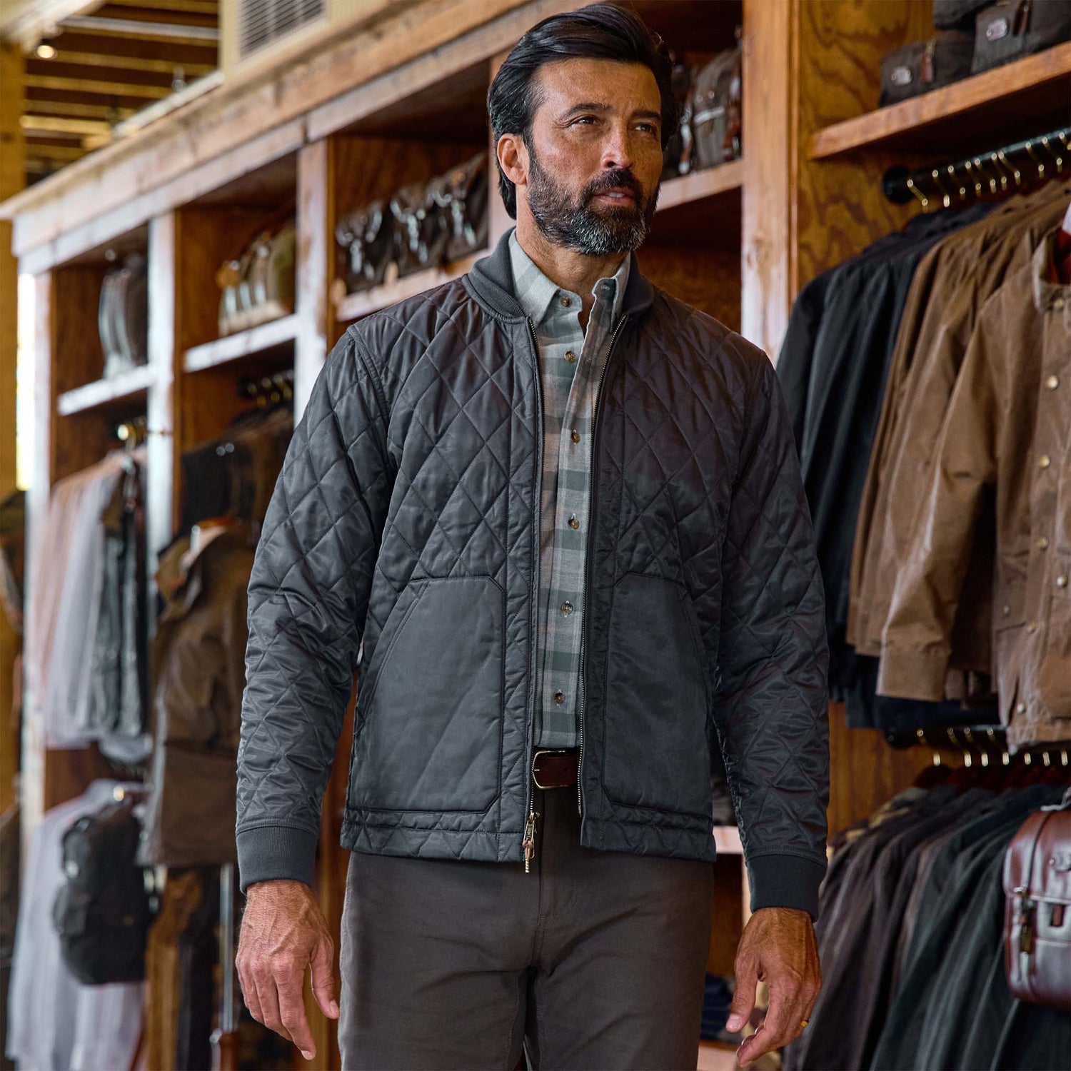A man in a Tom Beckbe Logan Bomber Jacket stands in a clothing store with jackets hanging on wooden racks.