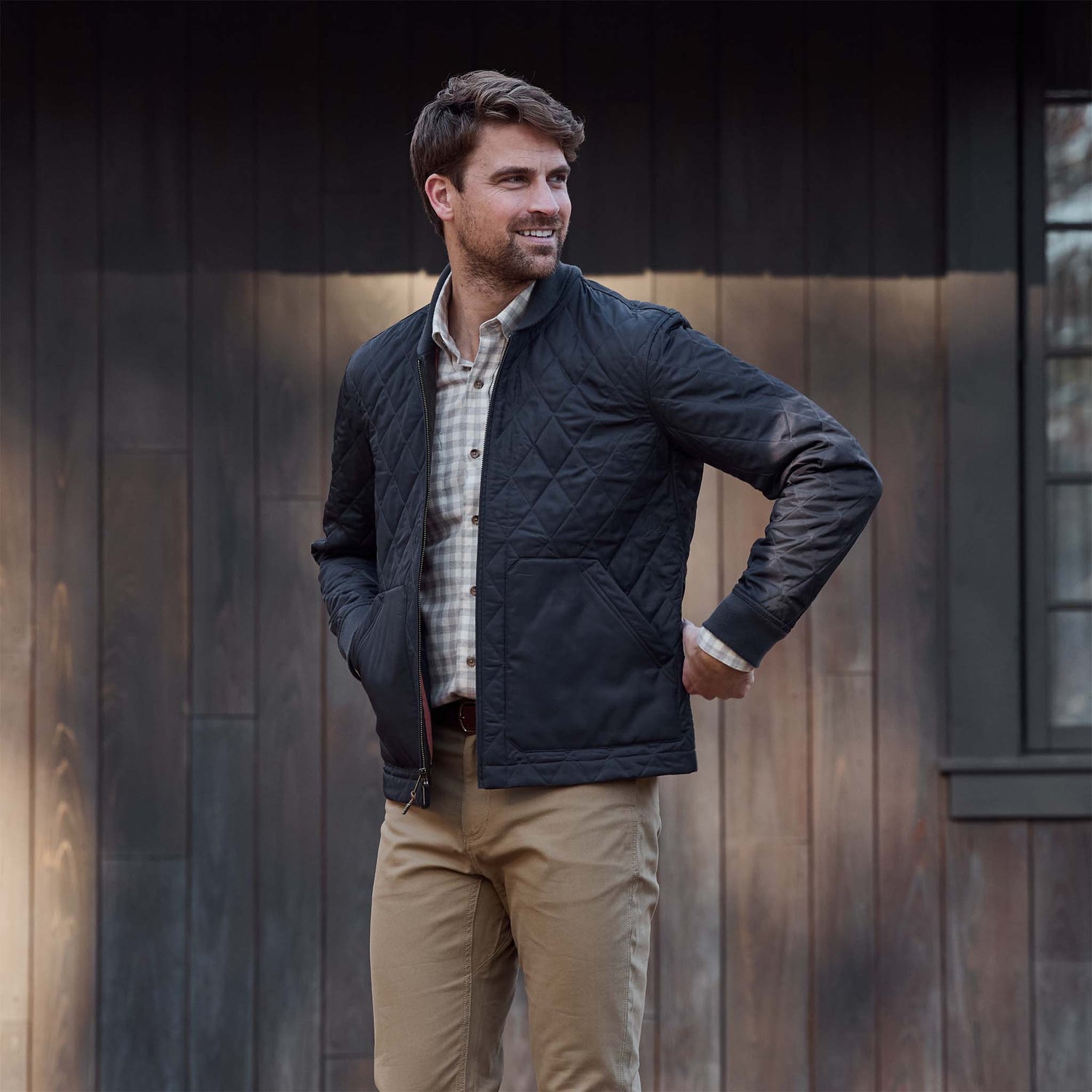 Man outdoors in a Tom Beckbe Logan Bomber Jacket, checked shirt, and khaki pants by a wooden building.