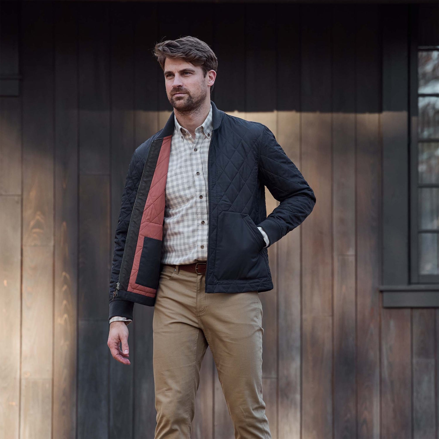Man in a checked shirt and khaki pants wears a Tom Beckbe Logan Bomber Jacket, standing outside by a wooden wall.