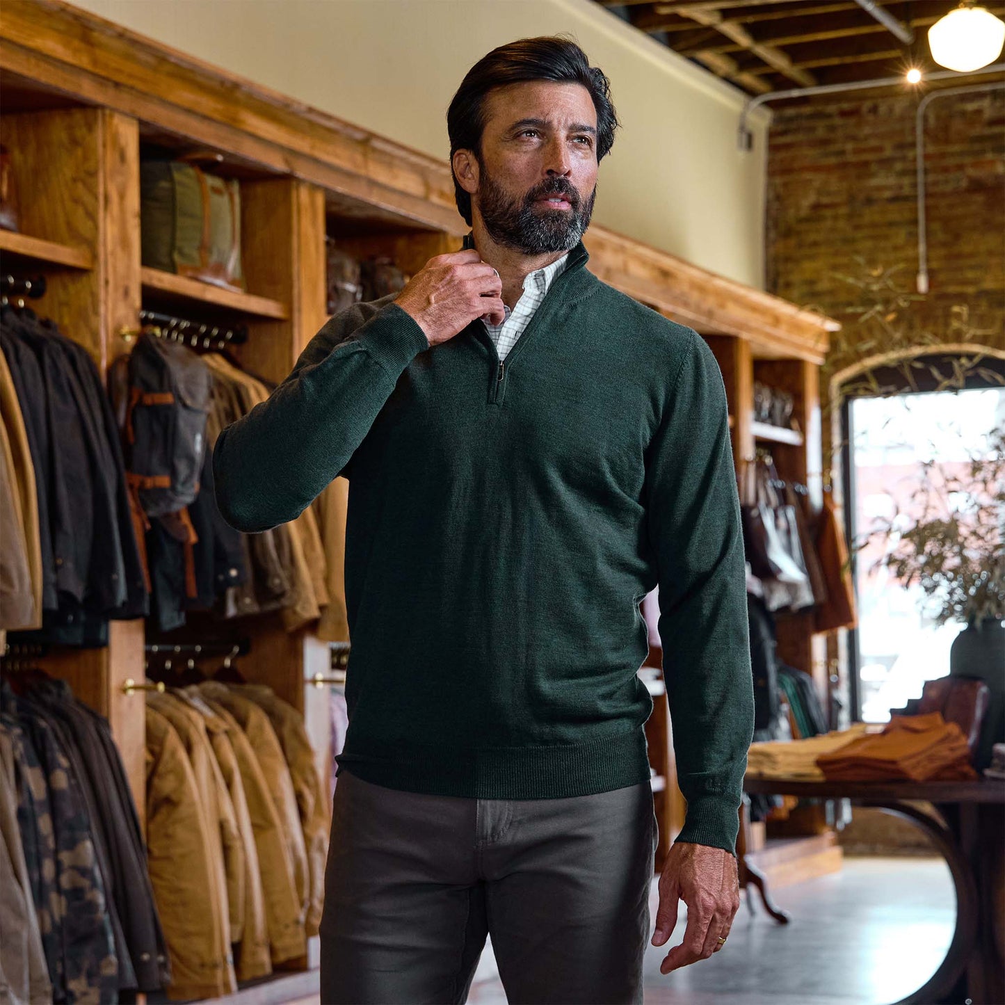 Man wearing a Tom Beckbe York Merino Quarter Zip in dark green and gray pants adjusts his collar in a clothing store.