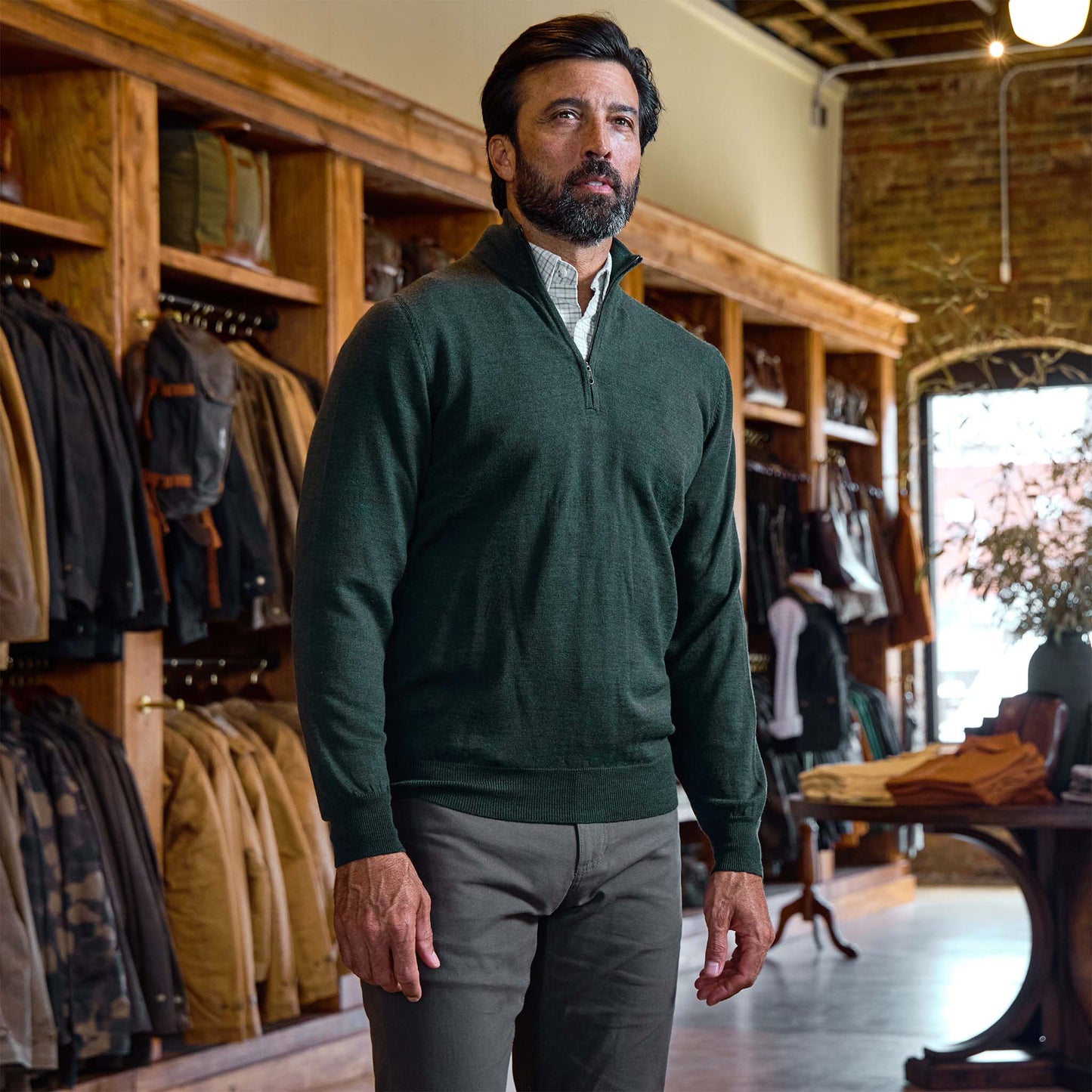 A man in a Tom Beckbe York Merino Quarter Zip sweater and gray pants stands in a store with wooden shelves and jackets.
