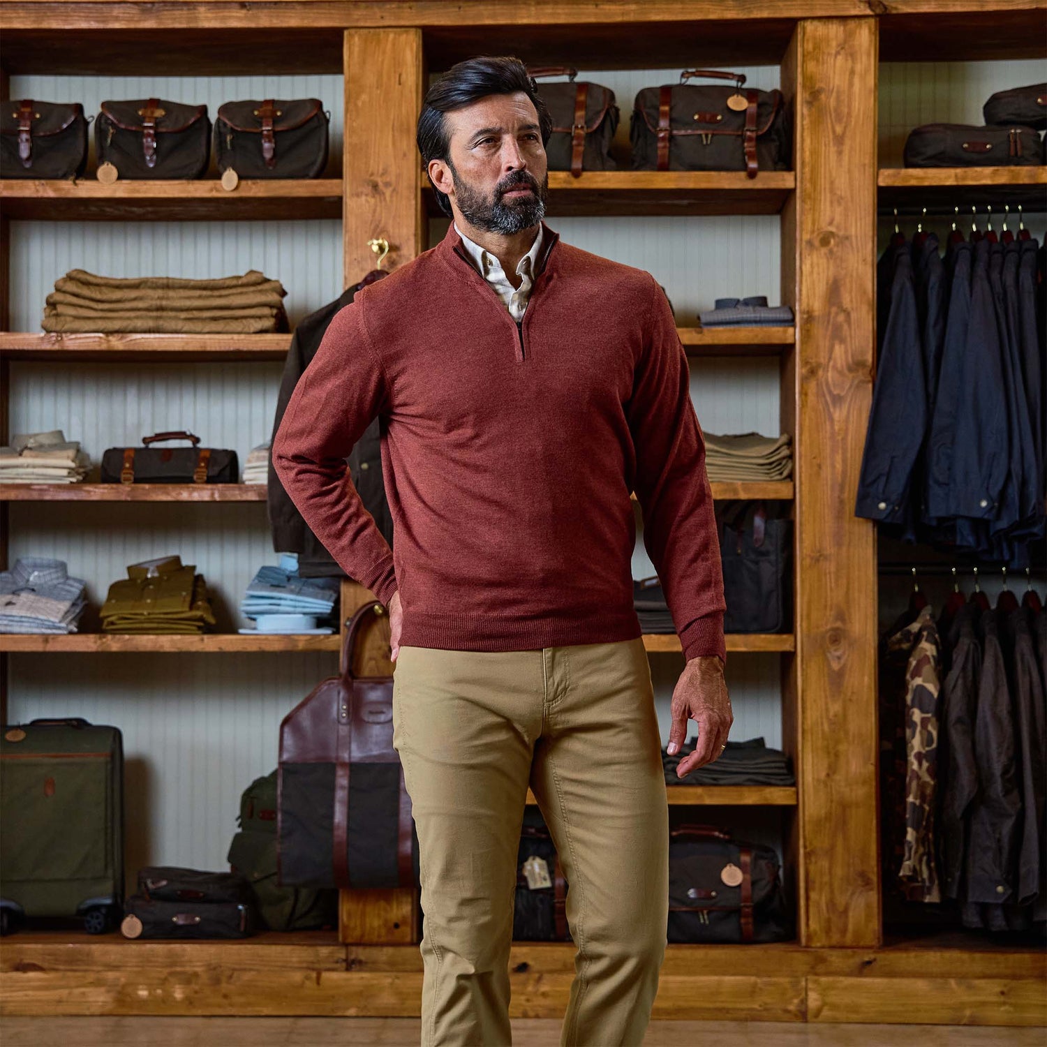 A man in a Tom Beckbe York Merino Quarter Zip and khaki pants stands in a store with shelves of clothes and bags behind him.