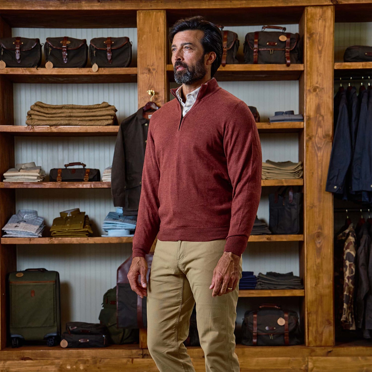 Man in a rust York Merino Quarter Zip by Tom Beckbe, made in Italy, stands among shelves and bags in a clothing store.