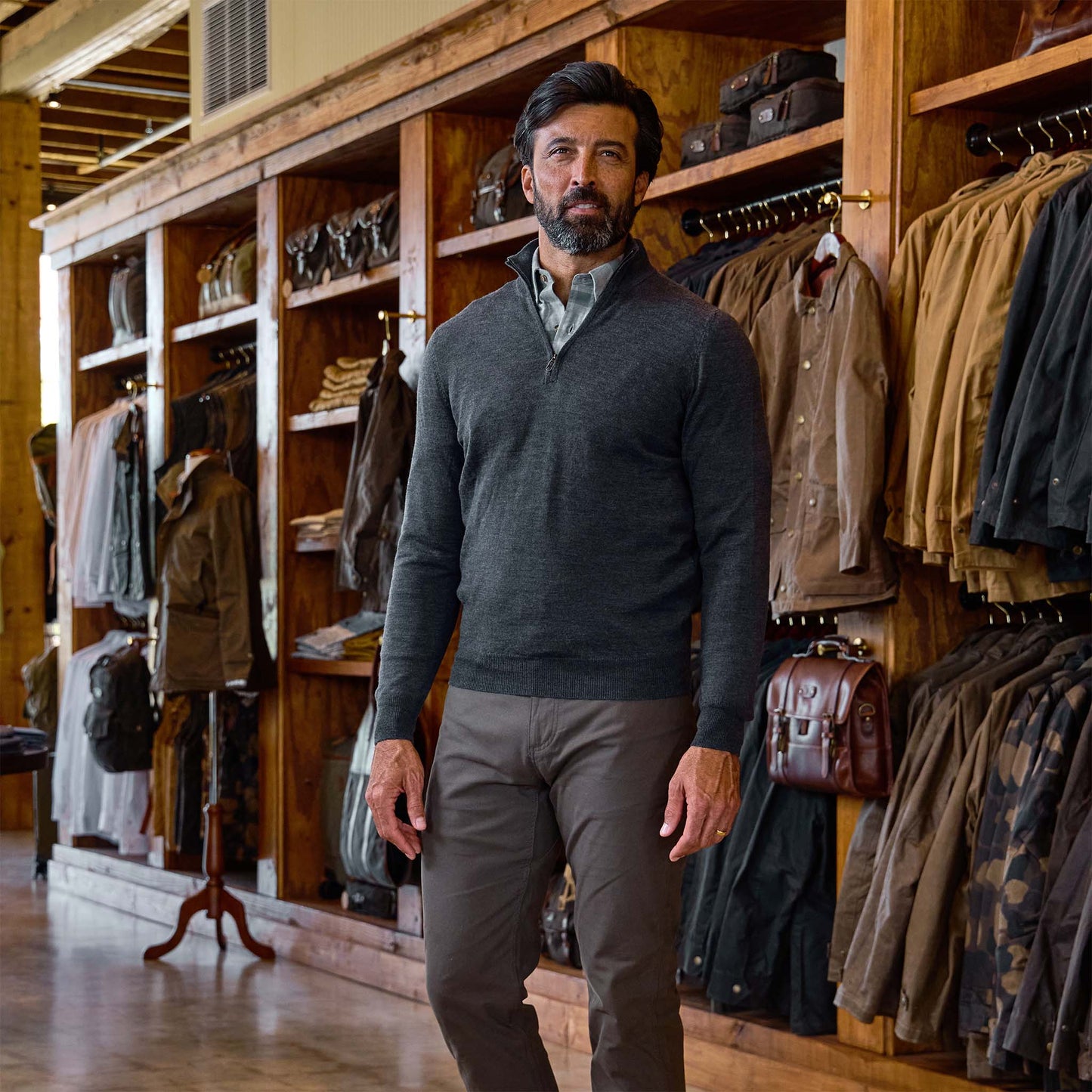 Man wearing Tom Beckbe York Merino Quarter Zip and brown pants stands in a store with wooden shelves and jackets.