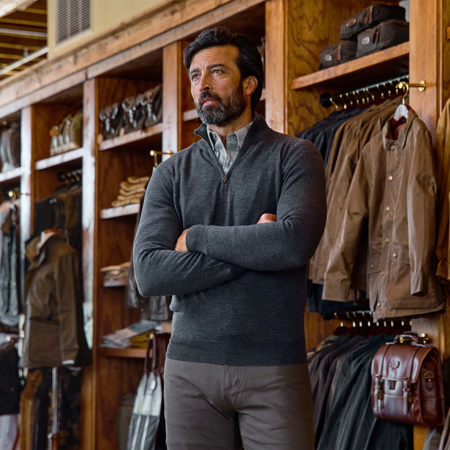 Man wearing the Tom Beckbe York Merino Quarter Zip stands with arms crossed in a store among jackets and bags.