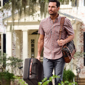A man carries a suitcase and Tom Beckbe Messenger Bag in front of a brick house with greenery.