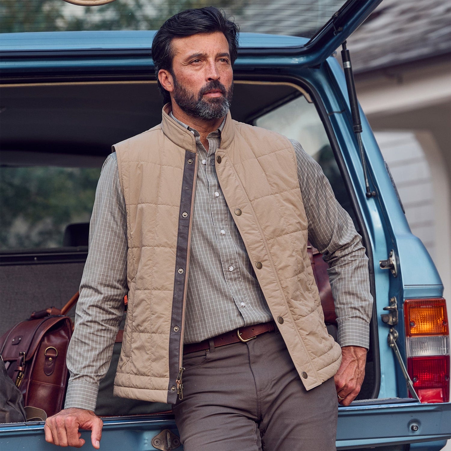 A man in a Monroe Poplin Shirt by Tom Beckbe leans against a blue SUV with travel bags in the back.