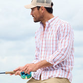 Man in a Tom Beckbe Monroe Poplin Shirt and cap fishing with a rod against a cloudy sky backdrop.