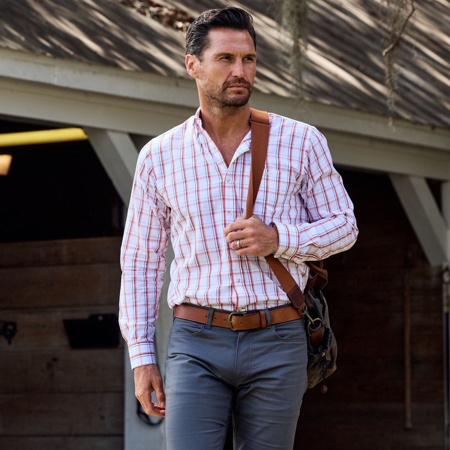 Man in a Tom Beckbe Monroe Poplin Shirt and gray pants holds a brown bag outdoors with a wooden building behind him.