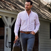 A man in a Monroe Poplin Shirt and gray pants holds a bag, strolling outside a white building with wooden beams.