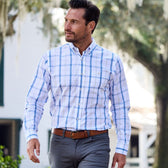 A man in a Monroe Poplin Shirt by Tom Beckbe walks outdoors, glancing sideways with trees and a building nearby.