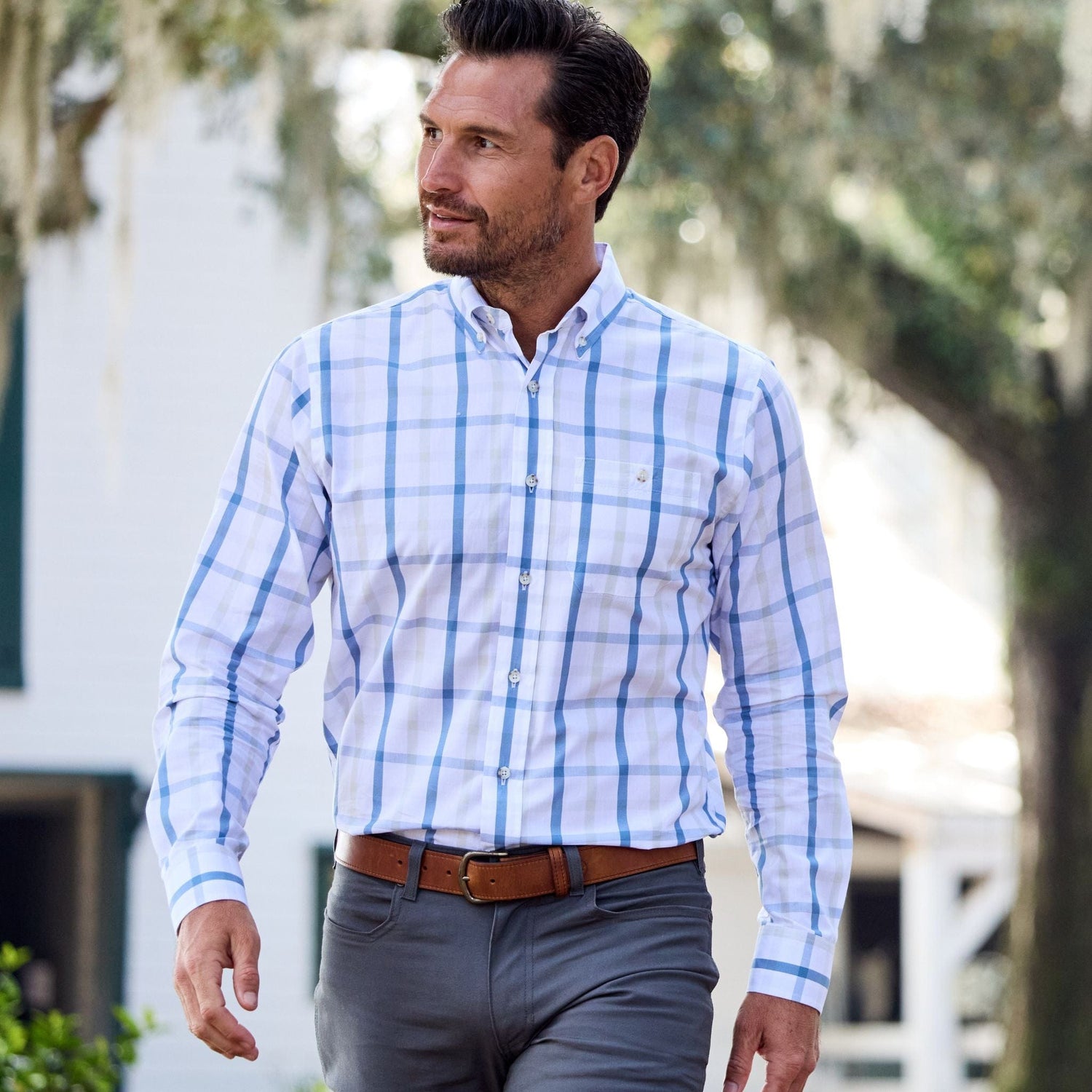 A man in a Monroe Poplin Shirt by Tom Beckbe walks outdoors, glancing sideways with trees and a building nearby.