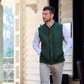A man outdoors in a green quilted vest and Tom Beckbes Monroe Poplin Shirt, hands in pockets, glances sideways.