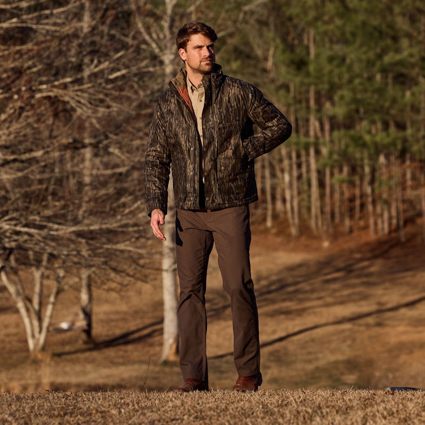 A person in a Tom Beckbe Fowler Jacket, Mossy Oak Original Bottomland, and brown pants stands on grass with trees behind.