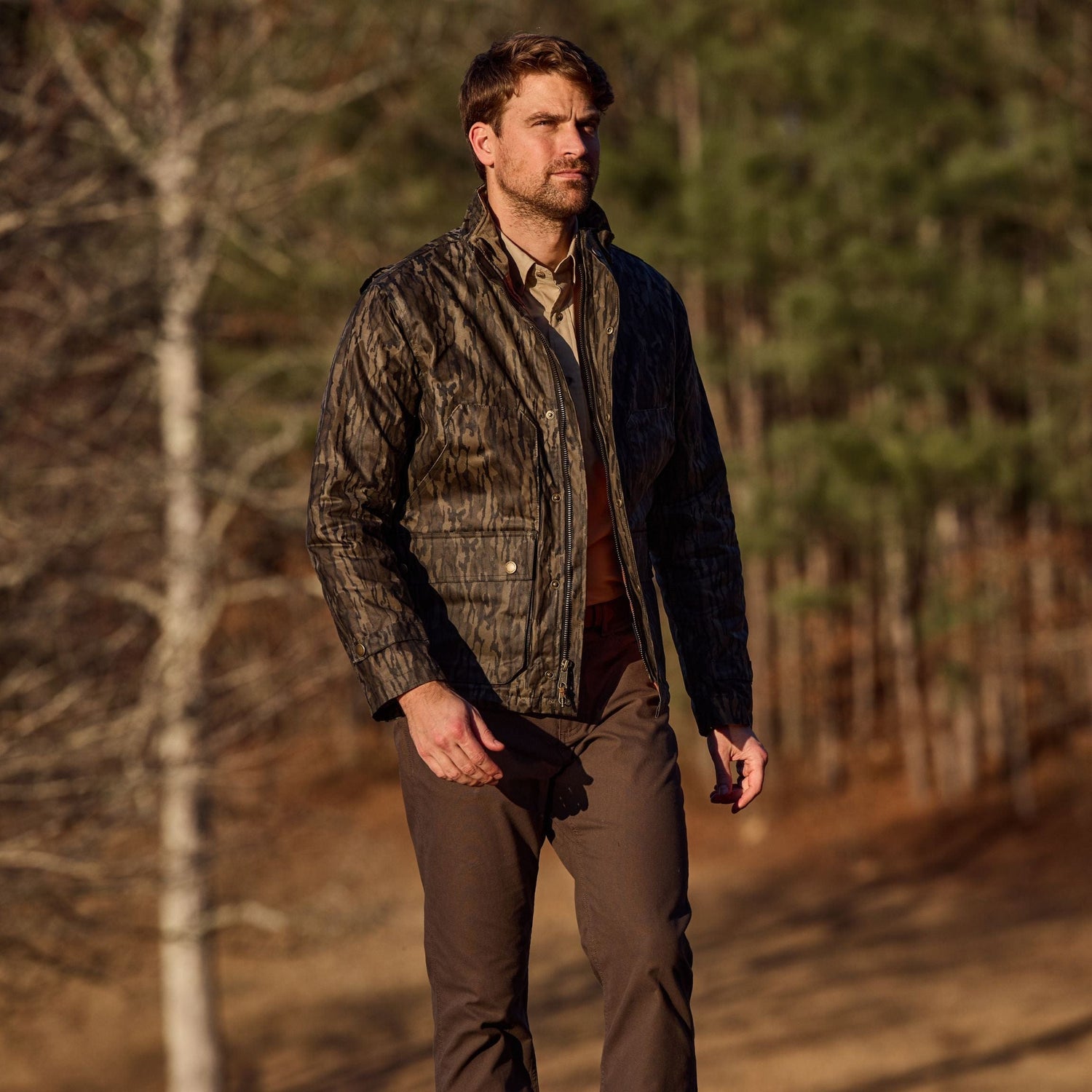 Man outdoors in a Tom Beckbe Fowler Jacket, Mossy Oak Original Bottomland, with brown pants; trees and grass in the background.