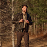 A man in a Tom Beckbe Fowler Jacket and brown pants walks outdoors near trees on a sunny day.