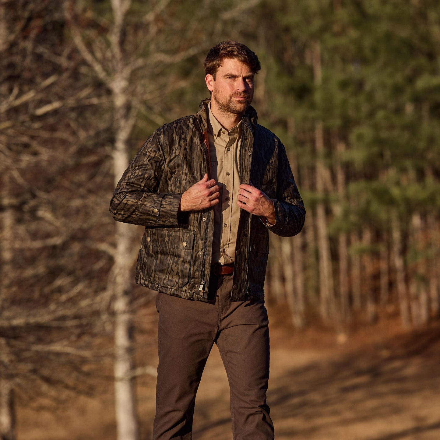 A man in a Tom Beckbe Fowler Jacket and brown pants walks outdoors near trees on a sunny day.