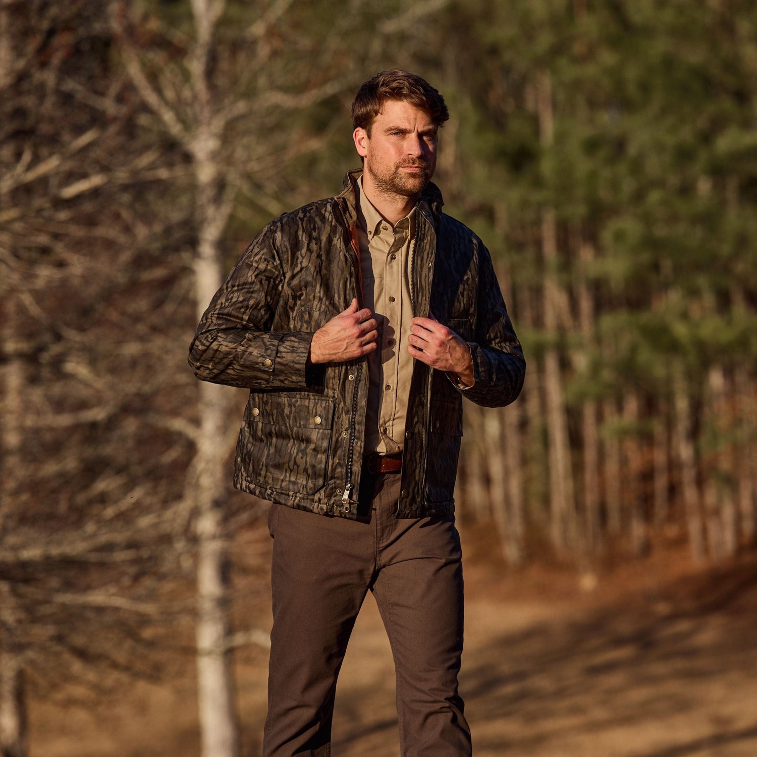 A man in a Tom Beckbe Fowler Jacket and brown pants walks outdoors near trees on a sunny day.