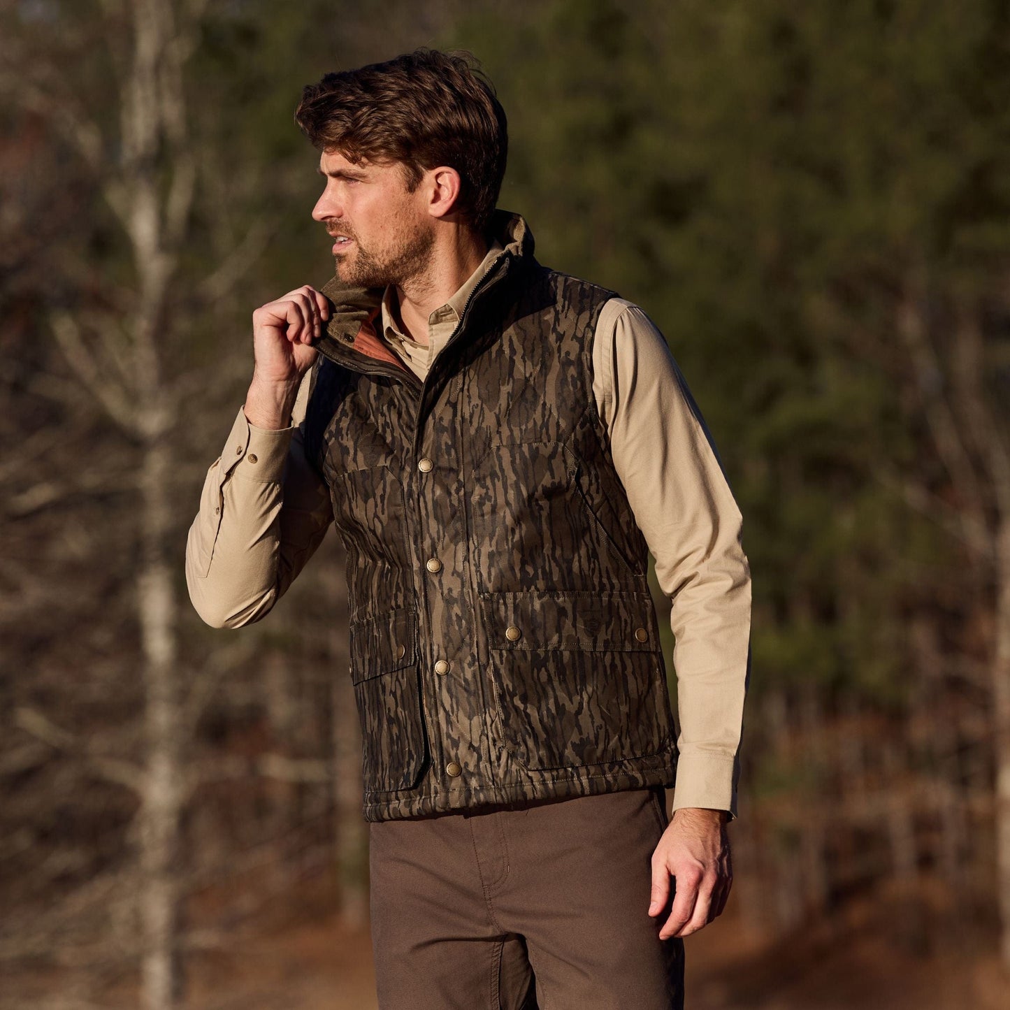 A man outdoors in a Tom Beckbe Mossy Oak Original Bottomland Fowler Vest adjusts his collar, with trees in the background.