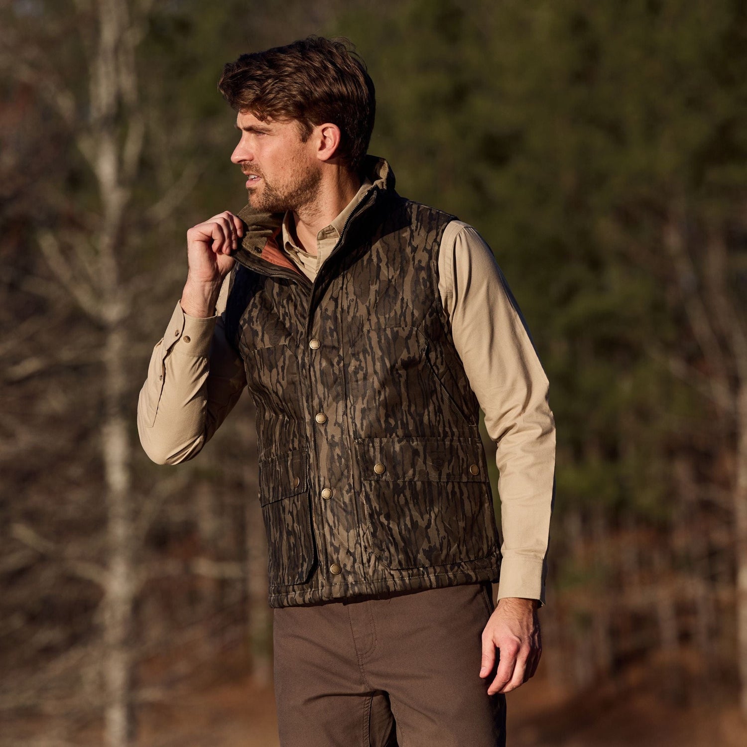 A man outdoors in a Tom Beckbe Mossy Oak Original Bottomland Fowler Vest adjusts his collar, with trees in the background.