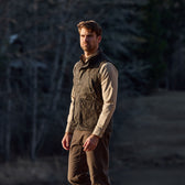 A man in a Tom Beckbe Kinsman Vest, Mossy Oak Original Bottomland, stands gazing sideways in a forest.