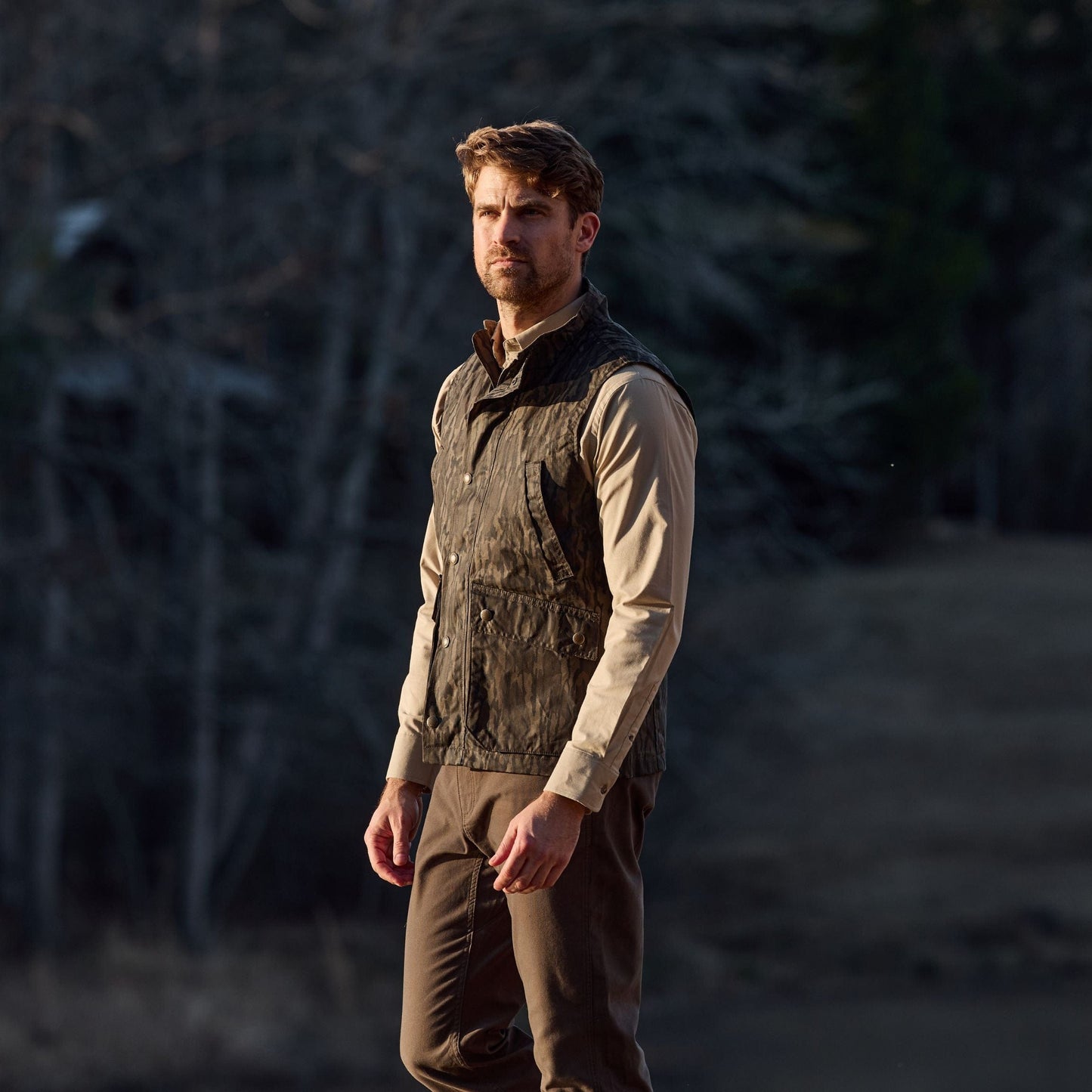 A man in a Tom Beckbe Kinsman Vest, Mossy Oak Original Bottomland, stands gazing sideways in a forest.
