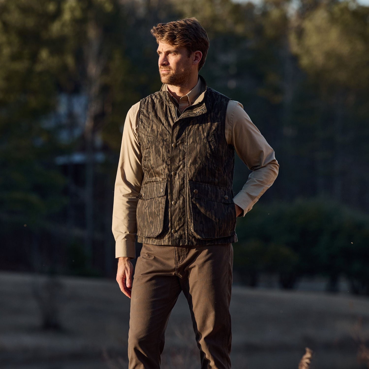 A man in a Tom Beckbe Kinsman Vest, Mossy Oak Original Bottomland, and beige shirt stands outdoors with trees behind.