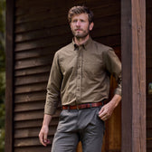 A bearded man leans against a beam, wearing a breathable Tom Beckbe Culver Oxford Shirt outside a wooden building.
