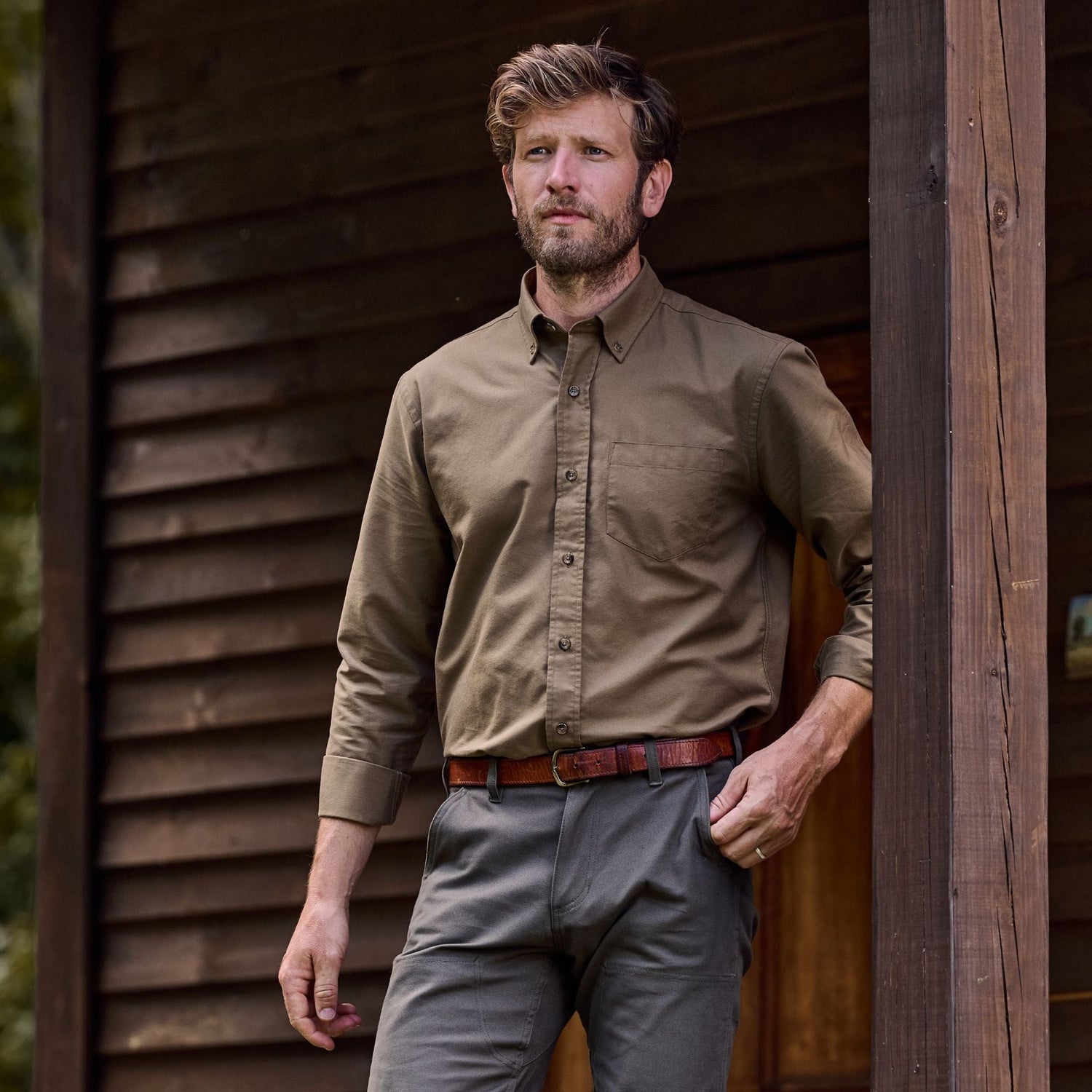 A bearded man leans against a beam, wearing a breathable Tom Beckbe Culver Oxford Shirt outside a wooden building.