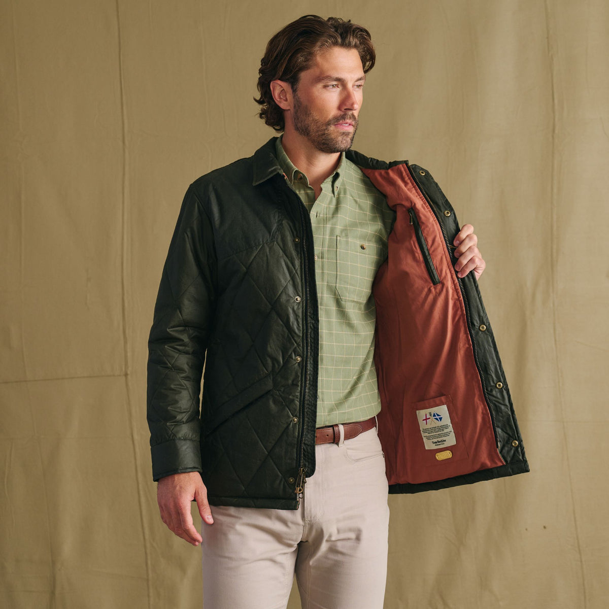 Man wears Tom Beckbe Paddock Quilted Jacket, holding it open to reveal its weatherproof lining and inner label.