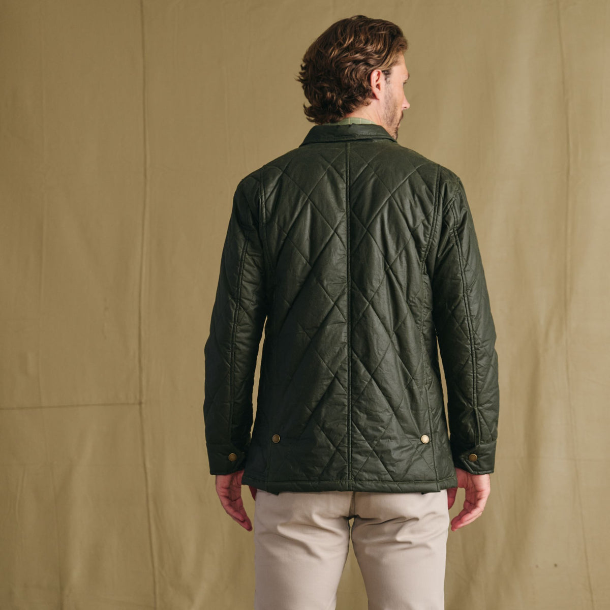 Man facing away, wearing Tom Beckbe Paddock Quilted Jacket in green and light pants, set against beige background.
