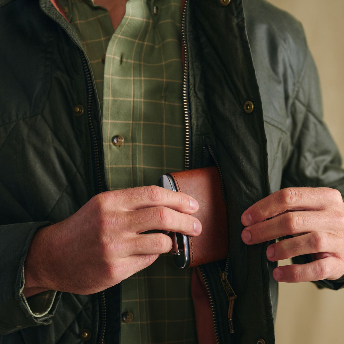 A person in a Tom Beckbe Paddock Quilted Jacket puts a brown leather wallet into the inner pocket of their coat.